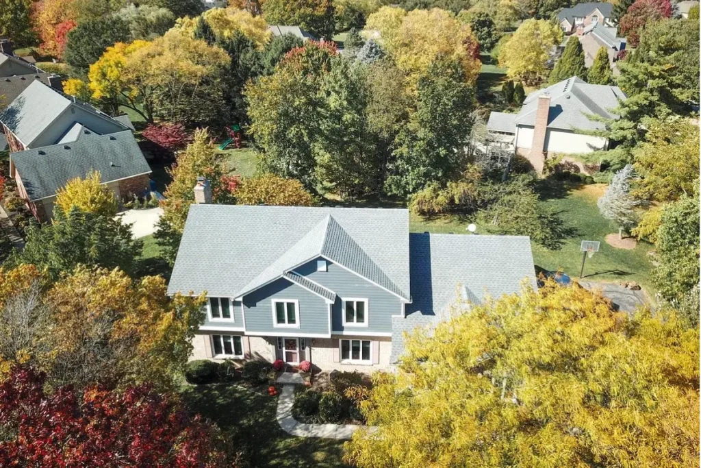 Aerial view of a suburban two-story house with a newly replaced gray shingle roof and blue exterior siding, surrounded by autumn trees.